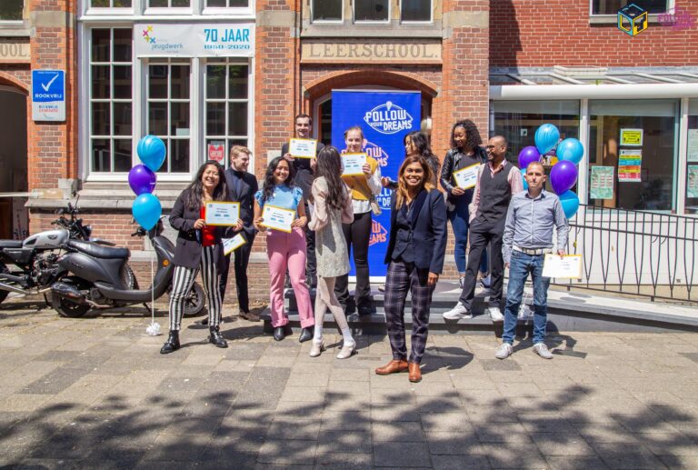 Wethouder Kavita Parbhudayal staat met een groep jongeren die diploma's vasthouden voor een versierde ingang van een gebouw.