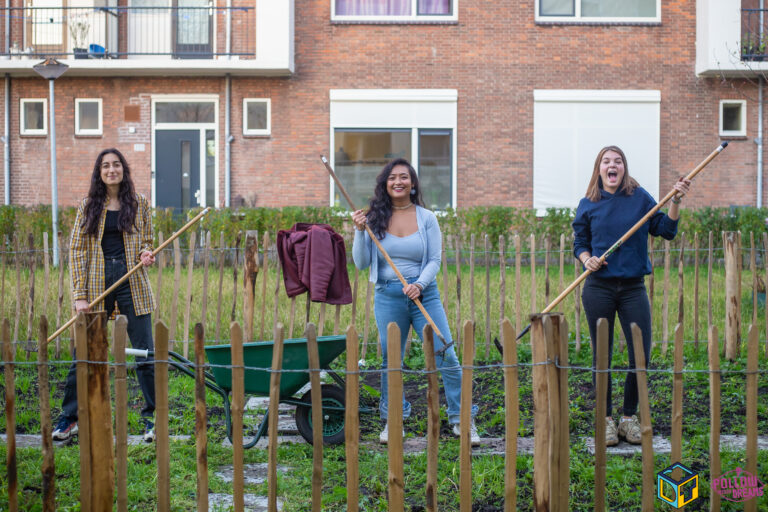 Drie jonge meiden poseren met harken en schoffels en een kruiwagen in een moestuin.